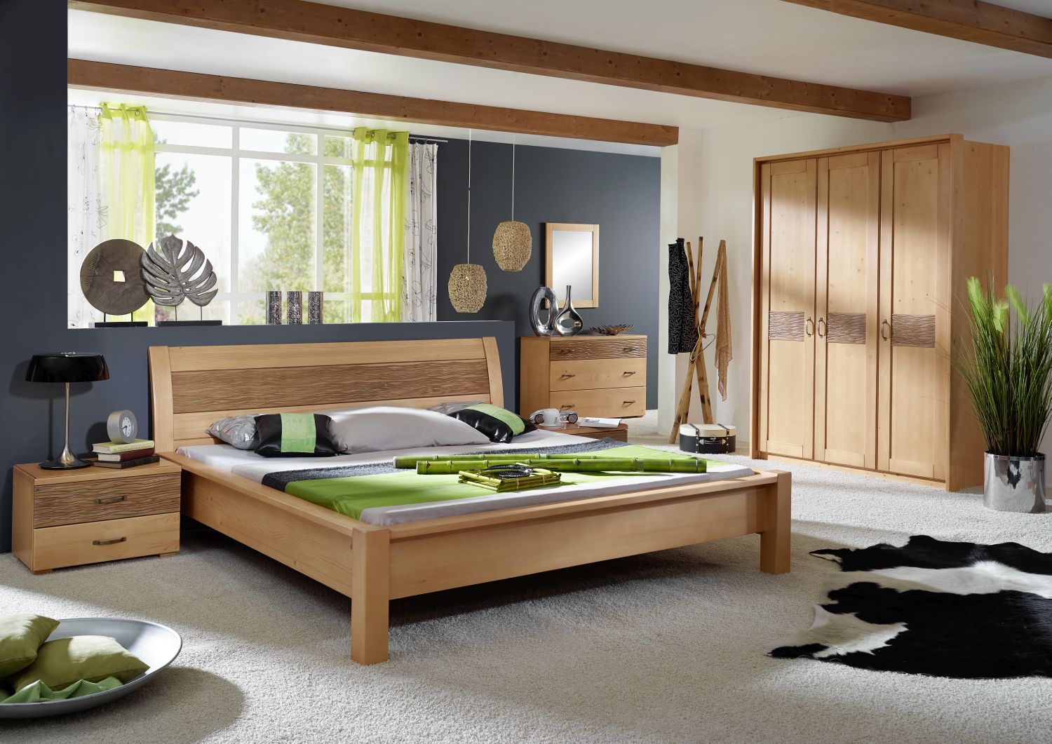 Modernes Schlafzimmer mit hellen Holzmöbeln, einem niedrigen Bett mit grünem und grauem Bettzeug, einem großen Kleiderschrank, einer Kommode und Wohndesign-Akzenten. Grüne Vorhänge und Pflanzen bringen Farbe ins Spiel; ein Kuhfellteppich liegt auf dem Teppichboden.
