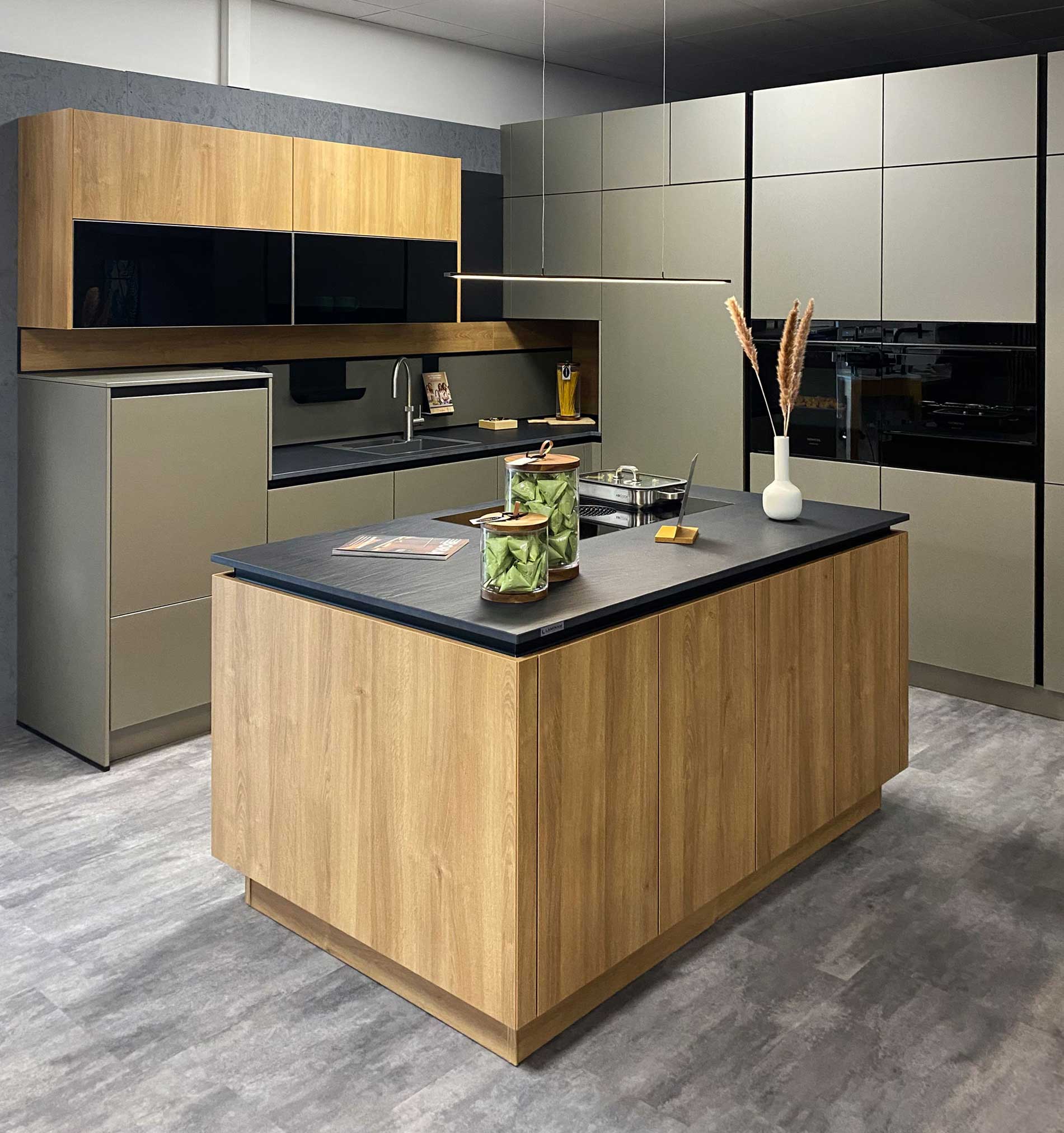 Eine moderne Küche mit einer Holzinsel mit schwarzer Arbeitsplatte, Induktionskochfeld, dekorativen Gläsern und einer Vase. Im Hintergrund unterstreichen Einbaugeräte, graue Schränke und ein Essbereich das Design.