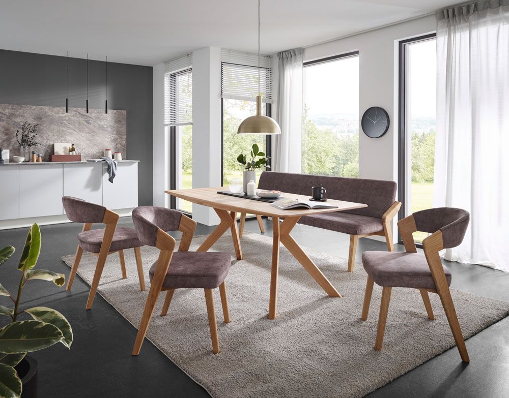 Modernes Esszimmer mit Holztisch, vier gepolsterten Stühlen, passender Bank, großen Fenstern, Pendelleuchte, Zimmerpflanze und weißen Schränken und einer grauen Akzentwand im Hintergrund.